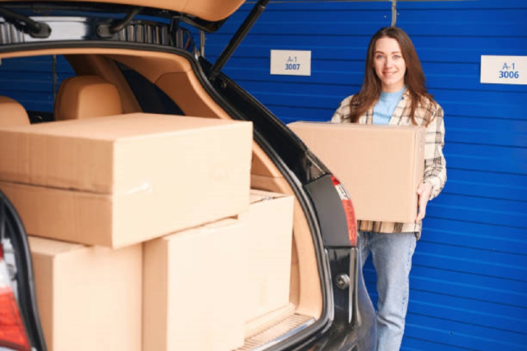 Young woman is standing near the open trunk of a car with boxes into warehouse and holding a big cardboard box